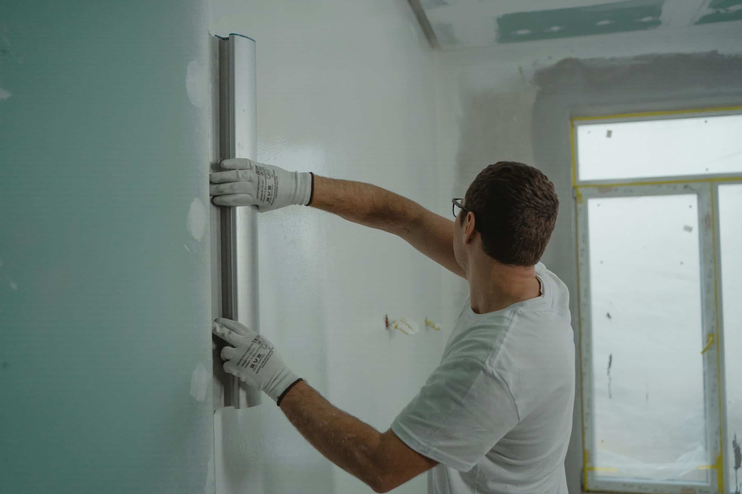 Optez pour une rénovation maitrisée. 3 A man in a white shirt and gloves using a metal ruler for indoor wall renovation work.
